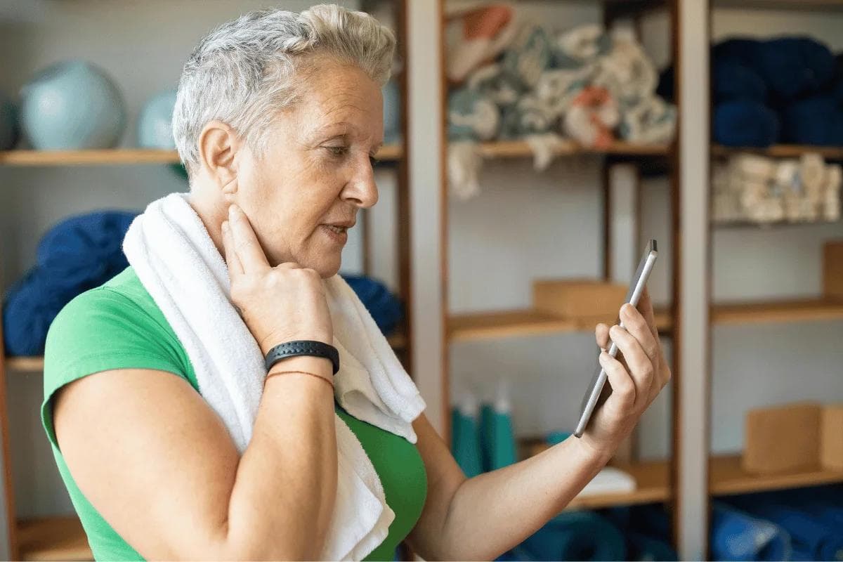 Senior woman with towel around neck looking at phone in fitness studio.