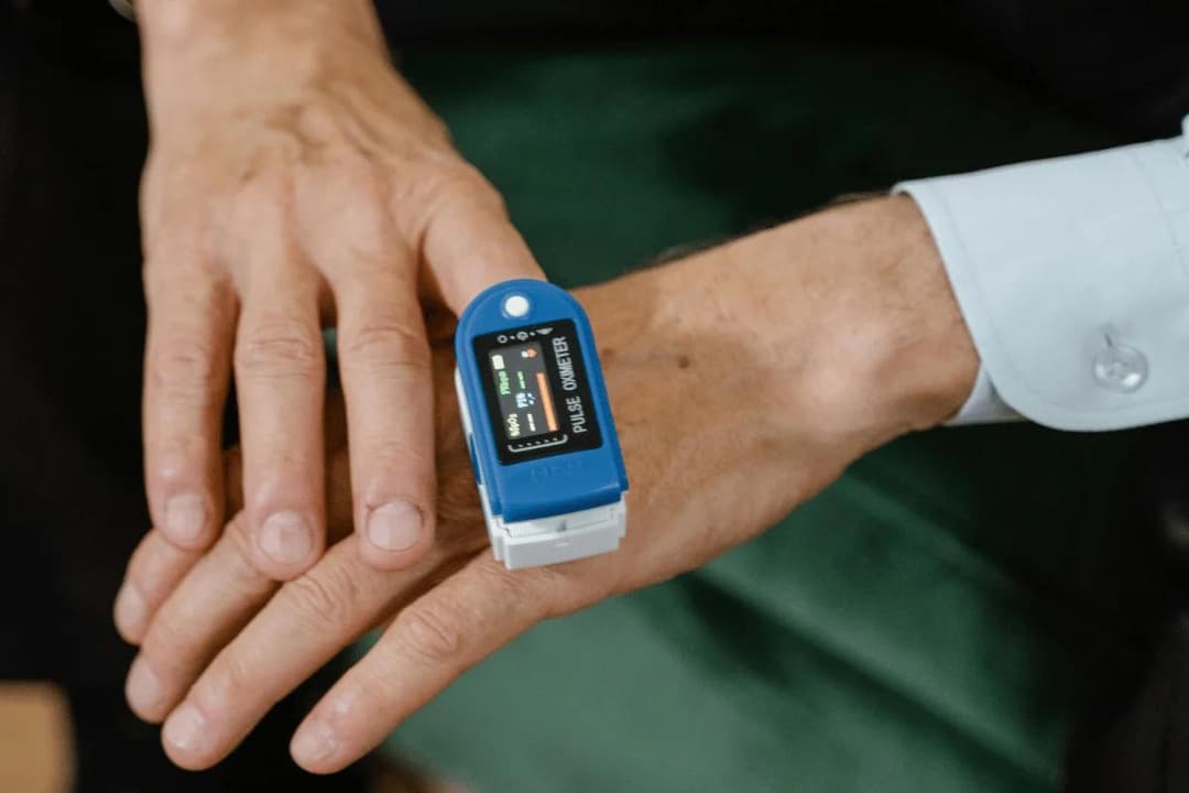A hand wearing a blue pulse oximeter on the index finger, displaying heart rate and oxygen levels, conveying a focus on health monitoring.