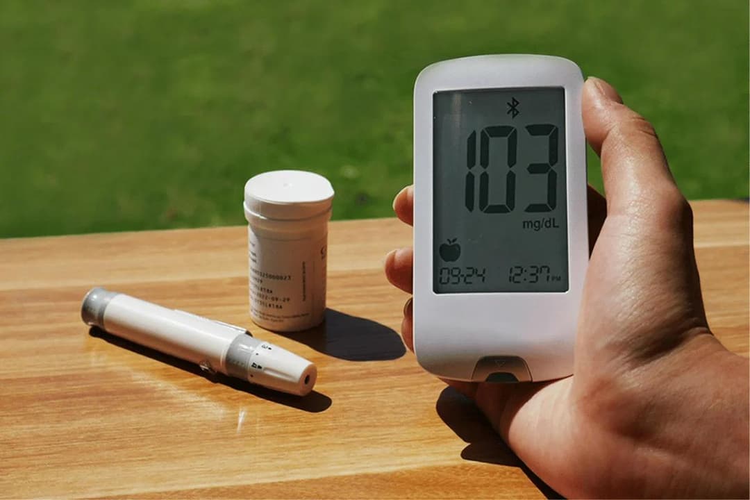 Hand holding glucose meter showing 103 mg/dL with test kit nearby.