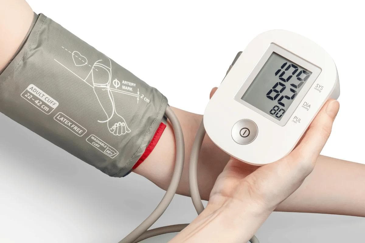 A hand holds a digital blood pressure monitor displaying readings while an arm with a gray cuff is positioned for measurement.
