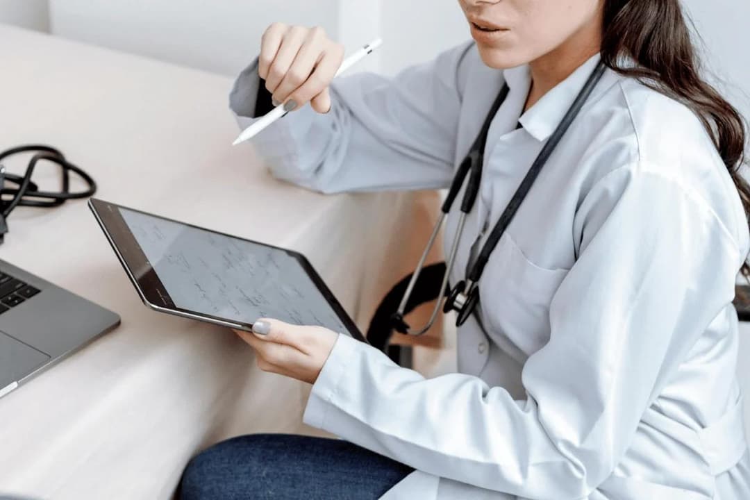 A healthcare professional in a white lab coat uses a tablet and pen for notes, with a stethoscope around their neck, sitting at a desk.