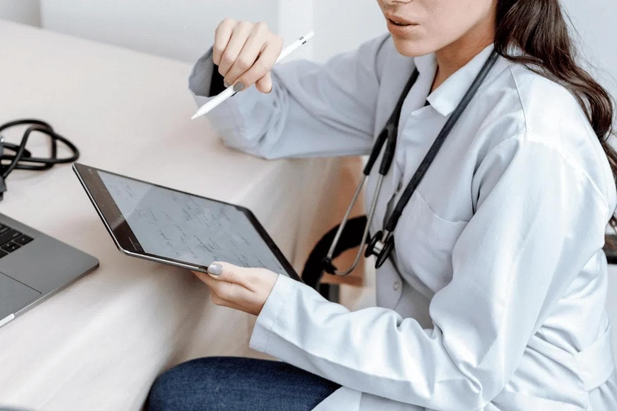 A healthcare professional in a white coat holds a tablet and a stylus, reviewing notes.