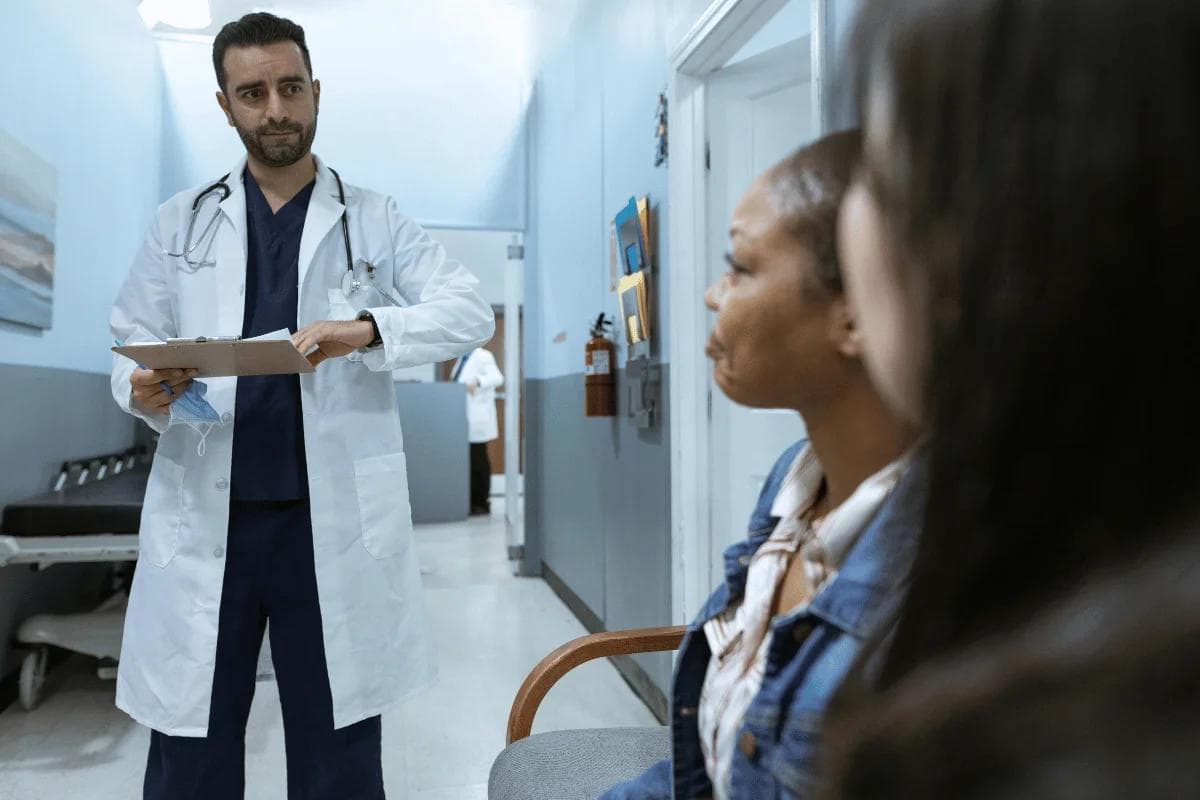 Doctor with stethoscope discusses patient care with two women in a hospital hallway, creating a professional setting.