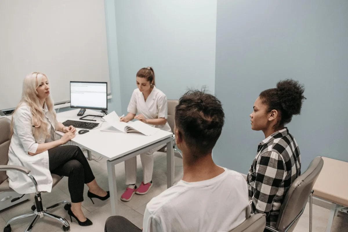 A female doctor and nurse, both in white coats, consult with a seated young man and woman in an office, creating a professional and attentive atmosphere.