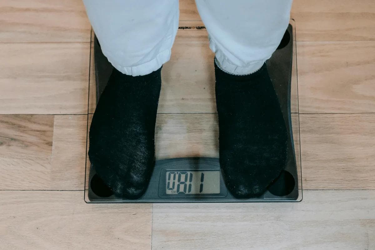 A person stands on a transparent digital bathroom scale, wearing white pants and black socks. The scale reads "00.1" on a light wooden floor.