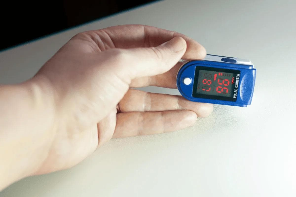A hand holding a blue digital pulse oximeter on a light surface. The display shows a blood oxygen level of 95% and a pulse rate of 87 BPM.