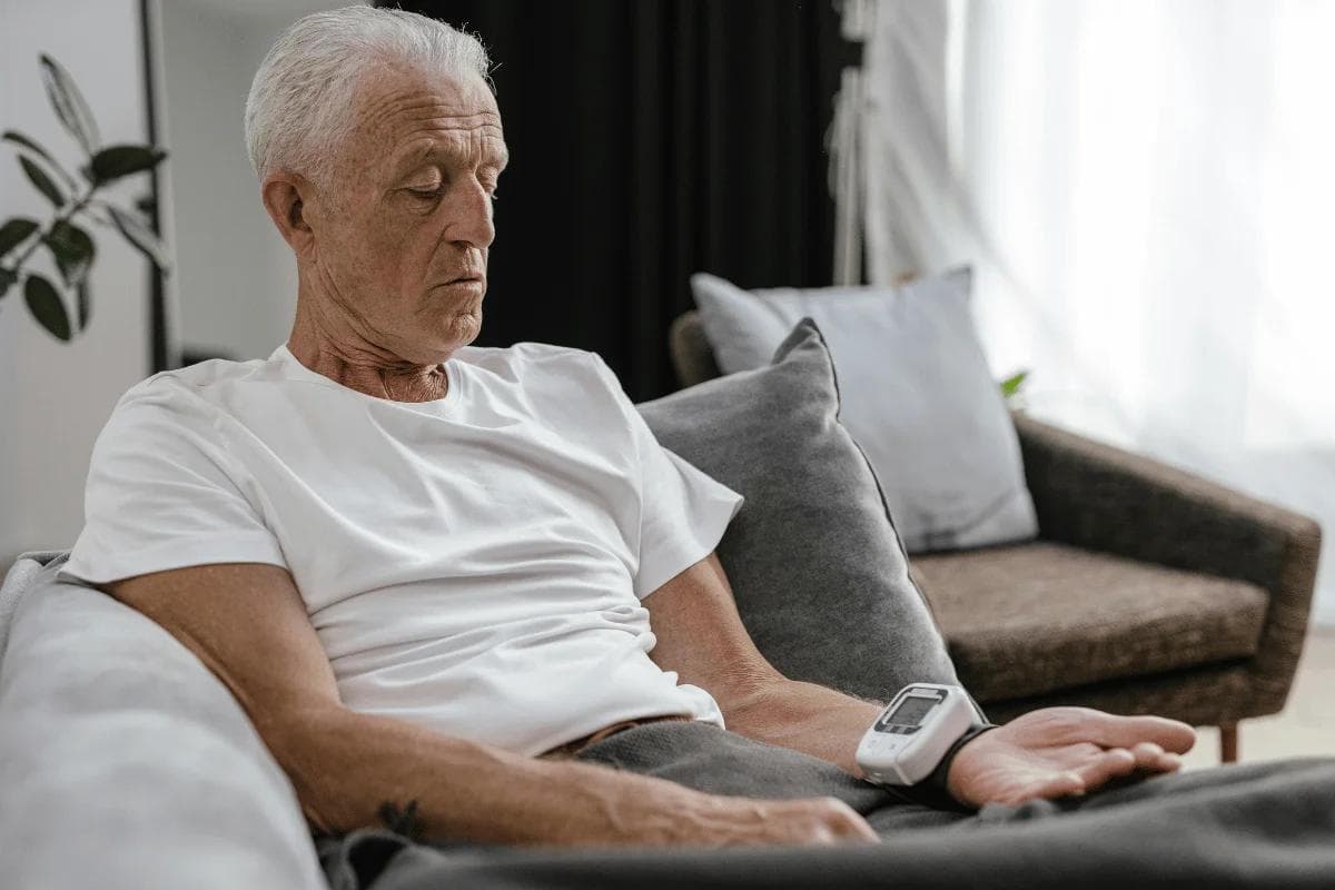 Elderly man with white hair, wearing a white t-shirt, sitting on a gray sofa, checking his blood pressure with a monitor, in a calm, softly lit room.