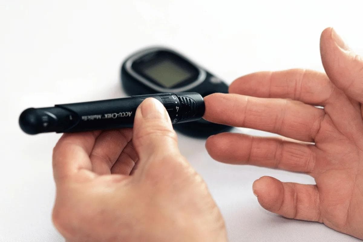 A person uses a black lancet device on their fingertip beside a glucose meter on a white surface. The scene conveys routine diabetes monitoring.