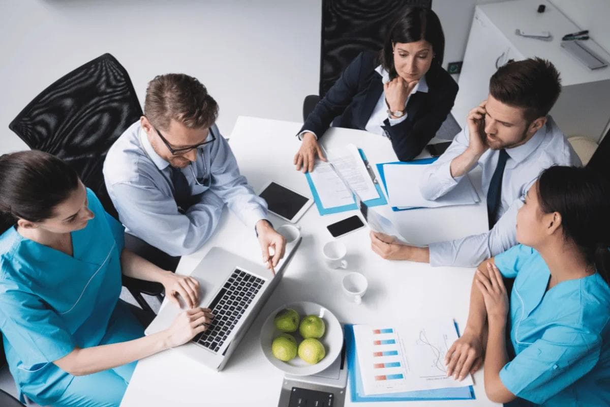 A diverse group of medical professionals and business people in a meeting around a table with a laptop, documents, and a bowl of green apples, appearing focused.