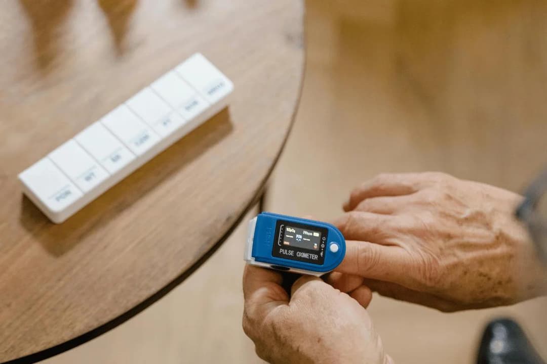 A person checks their pulse with a blue oximeter on their finger. Nearby, a pill organizer rests on a wooden table. The scene feels attentive and health-focused.