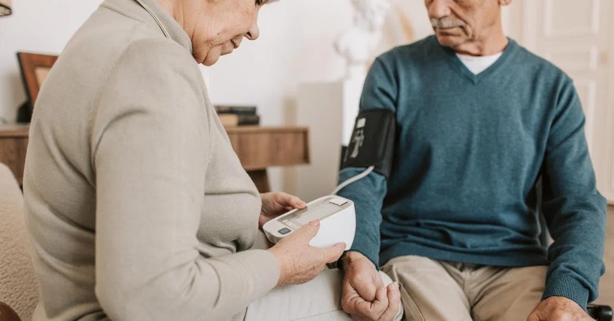An elderly woman measures an elderly man's blood pressure using a digital monitor.