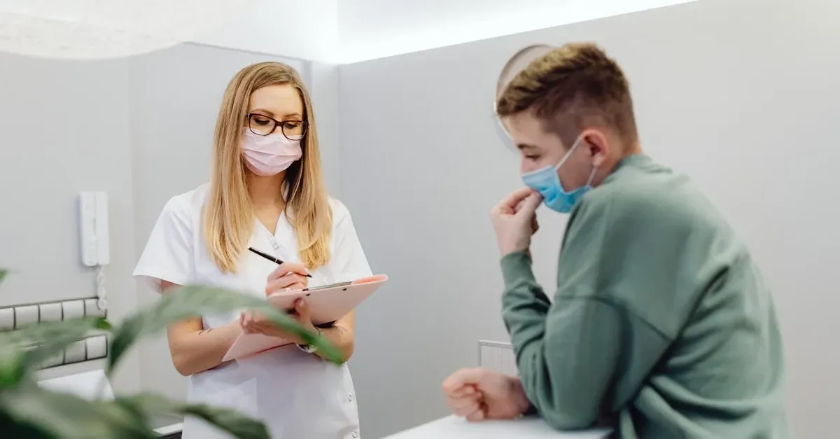 A female healthcare worker in a white uniform and pink mask writes on a clipboard while a young man in a green sweatshirt and blue mask watches.